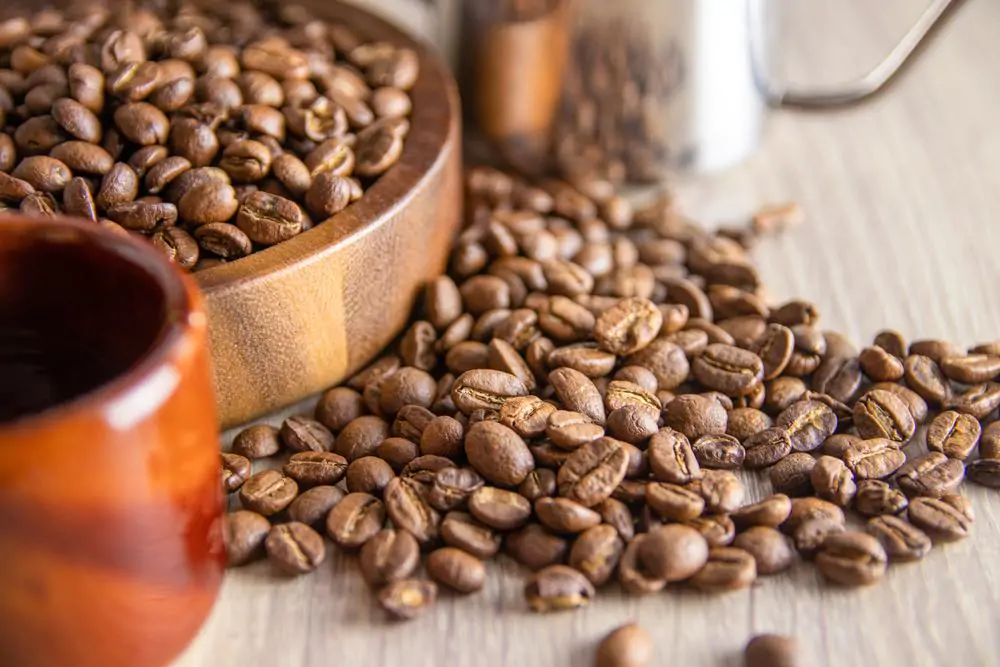 Some light roasted coffee beans grown in Thailand are on a wooden tray, some on the table top with a coffee mug on the left